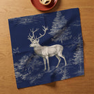 Navy Stag Woodland Christmas napkin opened face up on an oak table, showing a complete stag and woodland print beside a mince pie on a red plate