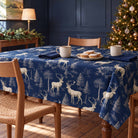 Navy Stag Woodland Christmas tablecloth draped over a dining table with cream mugs, mince pies and navy napkins in soft winter daylight.
