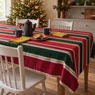 Red and green Signature Stripe Christmas tablecloth draped over a dining table with a dark coffee pot, two mugs, gold napkins and biscuits in a bright farmhouse room.