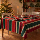 Red and green Signature Stripe Christmas tablecloth draped over a dining table with red napkins, pale green plates, candles and a small dish of walnuts.