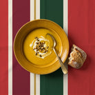 Signature Stripe Christmas tablecloth in red, green and burgundy seen from above, with a mustard bowl of parsnip soup, a brass spoon and a hunk of crusty bread