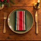 Red, green and burgundy Signature Stripe cotton napkin neatly folded on a dinner plate with cutlery and candlelight on a wooden Christmas table.