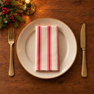 Red on ivory Ticking Stripe cotton napkin neatly folded on a dinner plate with cutlery and candlelight on a wooden Christmas table.