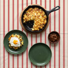 Ticking Stripe Christmas tablecloth in red on ivory seen from above, set for Boxing Day lunch with a cast-iron pan of bubble and squeak, two green plates, one with a fried egg, and a wooden bowl of chutney