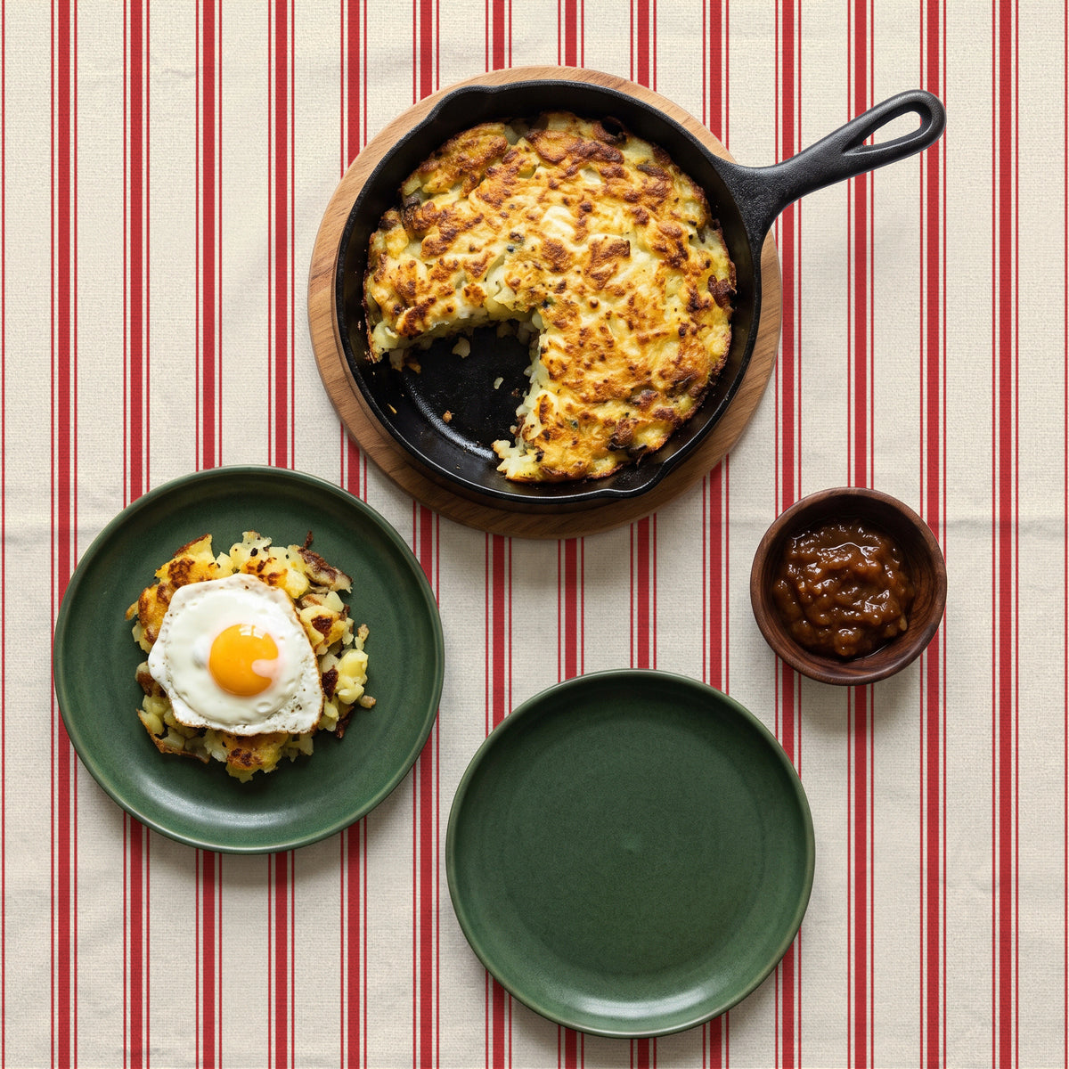 Ticking Stripe Christmas tablecloth in red on ivory seen from above, set for Boxing Day lunch with a cast-iron pan of bubble and squeak, two green plates, one with a fried egg, and a wooden bowl of chutney