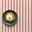 Red-on-ivory Ticking Stripe Christmas tablecloth seen from above, styled with one green plate of bubble and squeak and a fried egg in a closer composition.