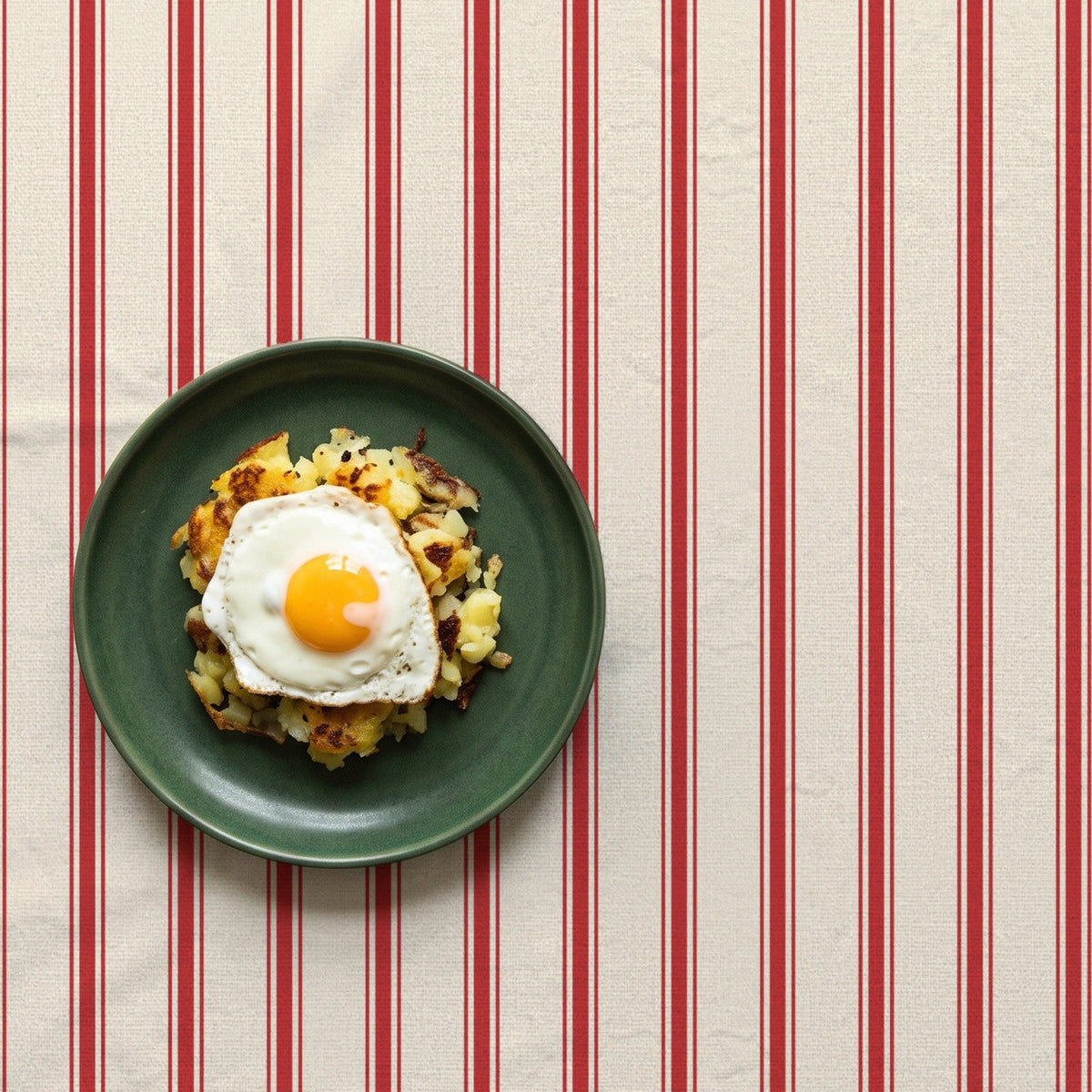 Red-on-ivory Ticking Stripe Christmas tablecloth seen from above, styled with one green plate of bubble and squeak and a fried egg in a closer composition.