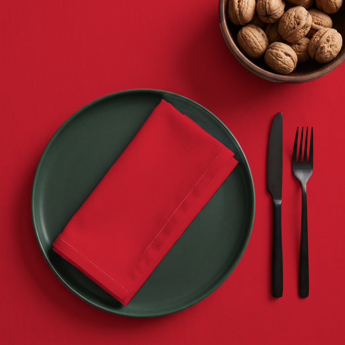 Red cotton Christmas napkin folded into an oblong on a green plate over the matching red tablecloth, with black cutlery and a bowl of walnuts.