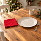 Red Plain cotton napkin in a compact square fold beside a dinner plate and coffee cup on a wooden table in daylight.