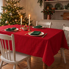 Red Plain Christmas tablecloth draped over a dining table with green napkins, ivory plates, walnuts and candles in a cream farmhouse room.