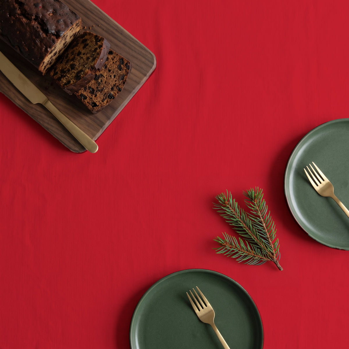 Plain red cotton Christmas tablecloth seen from above, with a sliced fruit loaf and a brass cake knife on a walnut board, two green side plates with brass forks and a sprig of fir.